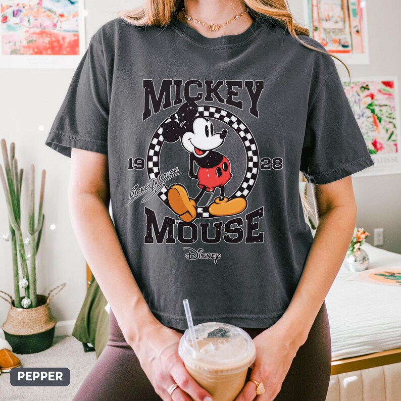 May include: A dark grey t-shirt with a Mickey Mouse graphic. The design includes the words "Mickey Mouse" and "Disney", the years "1928" and a chequered circle. The t-shirt has a relaxed fit.