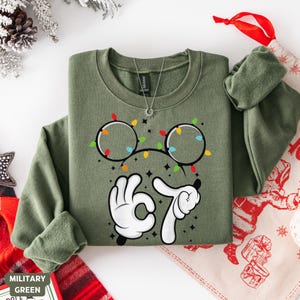 May include: Military green sweatshirt with a graphic of Mickey Mouse ears decorated with Christmas lights and the number 67 in white. The sweatshirt has long sleeves and a crew neck. A silver necklace is visible.