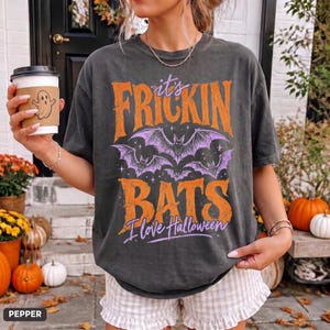 Op de afbeelding: Een antracietgrijs T-shirt met korte mouwen en de oranje en paarse tekst "it's FRICKIN BATS I love Halloween", voorzien van een afbeelding van drie paarse vleermuizen en kleine sterren. Het shirt wordt gedragen met een lichtgekleurde ruitjesbroek.