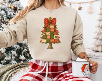 Può includere: Felpa beige con un design ad albero di Natale. L'albero è composto da quadrati patchwork rossi, verdi e bianchi, sormontato da un fiocco rosso e una stella dorata. La felpa è abbinata a pantaloni da pigiama a quadri rossi e bianchi.