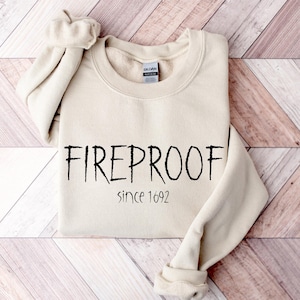 Könnte beinhalten: Ein cremefarbenes Sweatshirt mit dem Wort "FIREPROOF" in schwarzer, handschriftlicher Schrift. Darunter steht "since 1692". Das Sweatshirt liegt auf einer Holzoberfläche.