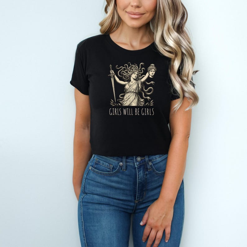 May include: Black t-shirt featuring a graphic of a woman with snakes for hair, holding a sword and a severed head. The text "GIRLS WILL BE GIRLS" is printed below the graphic. The shirt is worn by a person wearing blue jeans.