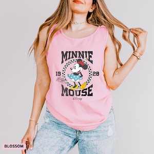 K&ouml;nnte beinhalten: Ein hellrosa Tanktop mit einem Minnie Mouse-Motiv. Das Design umfasst die schwarzen W&ouml;rter "MINNIE MOUSE", die Jahreszahlen "1928" und "19" sowie das Disney-Logo. Das Tanktop wird mit einer blauen Jeans getragen.