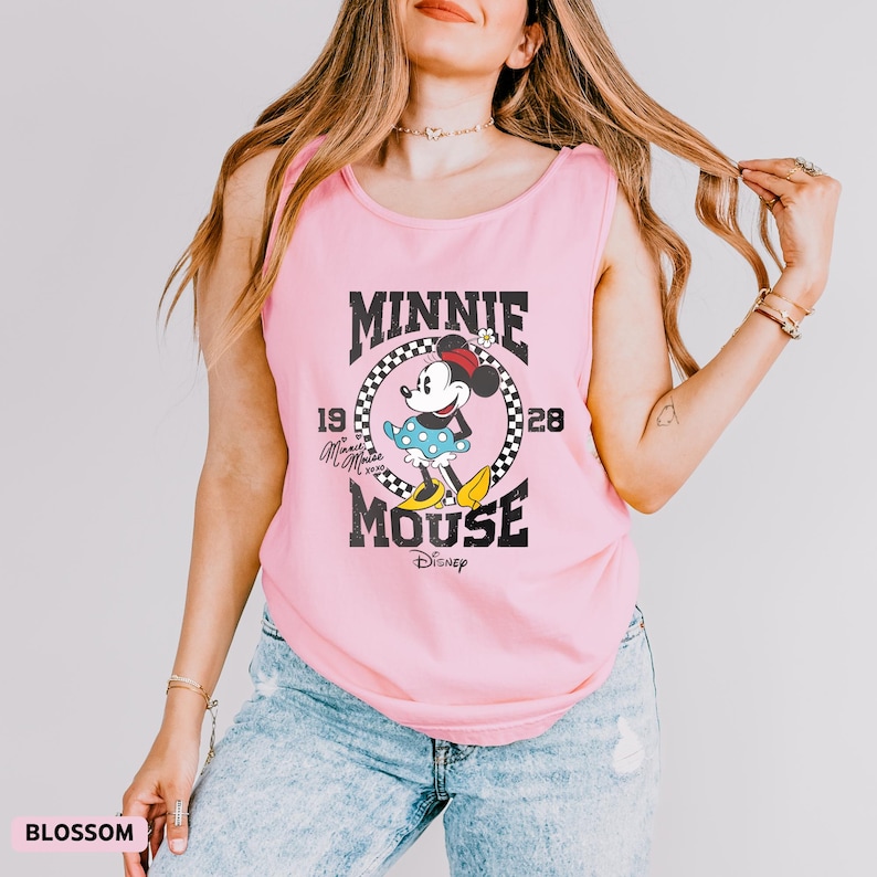 K&ouml;nnte beinhalten: Ein hellrosa Tanktop mit einem Minnie Mouse-Motiv. Das Design umfasst die schwarzen W&ouml;rter "MINNIE MOUSE", die Jahreszahlen "1928" und "19" sowie das Disney-Logo. Das Tanktop wird mit einer blauen Jeans getragen.