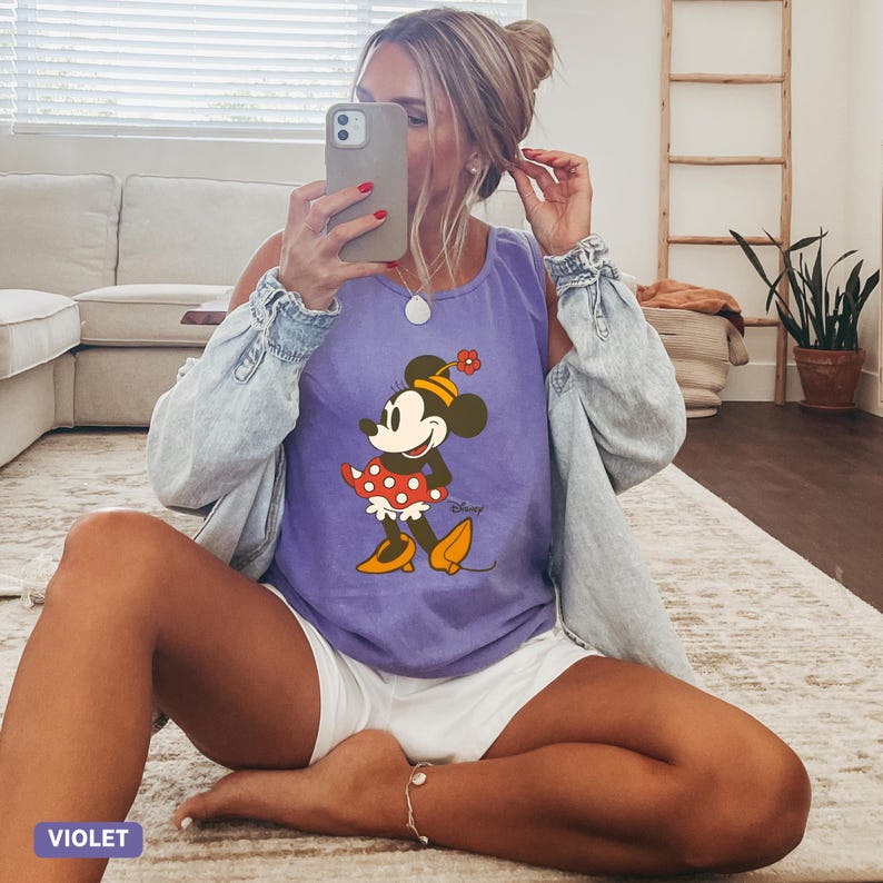 Puede incluir: Una camiseta sin mangas de color violeta con un estampado gr&aacute;fico de Minnie Mouse que lleva una falda de lunares rojos y blancos, un sombrero amarillo con una flor roja y zapatos amarillos. La camiseta tiene cuello redondo y se lleva con una camisa vaquera azul claro.
