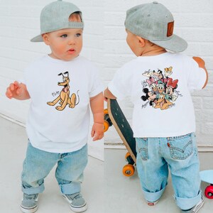 Pode incluir: Uma crian&ccedil;a pequena vestindo uma camiseta branca com Pluto na frente e personagens da Disney nas costas. A crian&ccedil;a tamb&eacute;m est&aacute; usando jeans azul claro, t&ecirc;nis cinza e um bon&eacute; de beisebol cinza. A crian&ccedil;a est&aacute; segurando um skate.