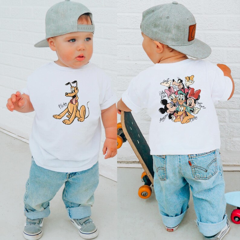 Pode incluir: Uma crian&ccedil;a pequena vestindo uma camiseta branca com Pluto na frente e personagens da Disney nas costas. A crian&ccedil;a tamb&eacute;m est&aacute; usando jeans azul claro, t&ecirc;nis cinza e um bon&eacute; de beisebol cinza. A crian&ccedil;a est&aacute; segurando um skate.