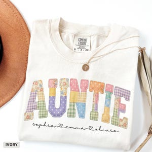 May include: Ivory-colored t-shirt with the word "AUNTIE" in patchwork letters. The names "sophia", "emma", and "olivia" are written below. A gold necklace and a brown hat are also visible.