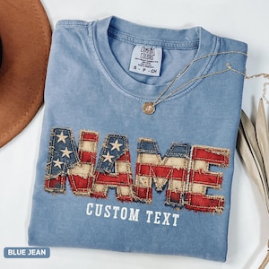 May include: A blue jean colored t-shirt with a Comfort Colors label at the neck. The shirt features the word NAME in a distressed American flag pattern with frayed edges, and the words CUSTOM TEXT printed in white below the large letters.