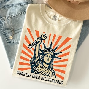 May include: Cream-coloured t-shirt with a graphic of the Statue of Liberty holding a wrench, set against orange sunburst rays. The text "WORKERS OVER BILLIONAIRES" is printed below. The shirt is folded on a surface with jeans and a hat.