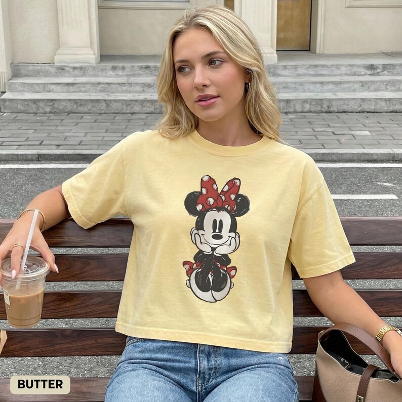 K&ouml;nnte beinhalten: Hellgelbes T-Shirt mit einem Minnie Mouse-Motiv, einschlie&szlig;lich einer roten Schleife mit wei&szlig;en Punkten. Das T-Shirt hat eine lockere Passform und kurze &Auml;rmel. Das Wort "BUTTER" ist unten links im Bild aufgedruckt.