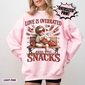 May include: Light pink sweatshirt with an oversized print. The graphic features a cherub wearing heart-shaped sunglasses, sitting on a bench and holding a bag of snacks. Text reads "LOVE IS OVERRATED" and "PASS THE SNACKS".