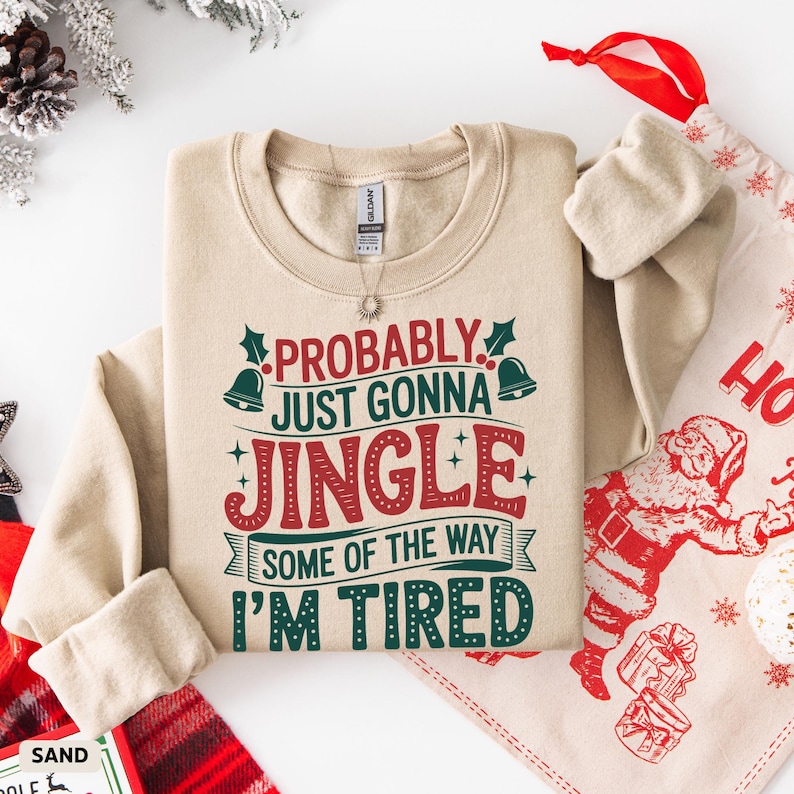 May include: A sand-colored sweatshirt with the text "PROBABLY JUST GONNA JINGLE SOME OF THE WAY I'M TIRED" in red and green. The sweatshirt is folded neatly, with a Christmas-themed design in the background.
