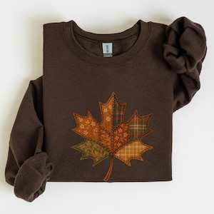 May include: A brown sweatshirt with a large appliquéd maple leaf design. The leaf is made from various patterned fabrics in orange, green, and brown. The sweatshirt has long sleeves and a crew neck.