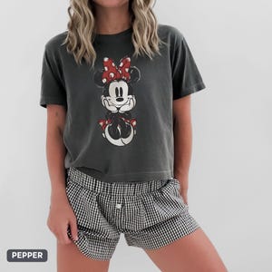 K&ouml;nnte beinhalten: Dunkelgraues T-Shirt mit einem verblassten Minnie Mouse-Motiv, roter Schleife und passenden Schuhen. Dazu eine schwarz-wei&szlig; karierte Shorts. Das Wort "PEPPER" steht in einem grauen Feld unten links.