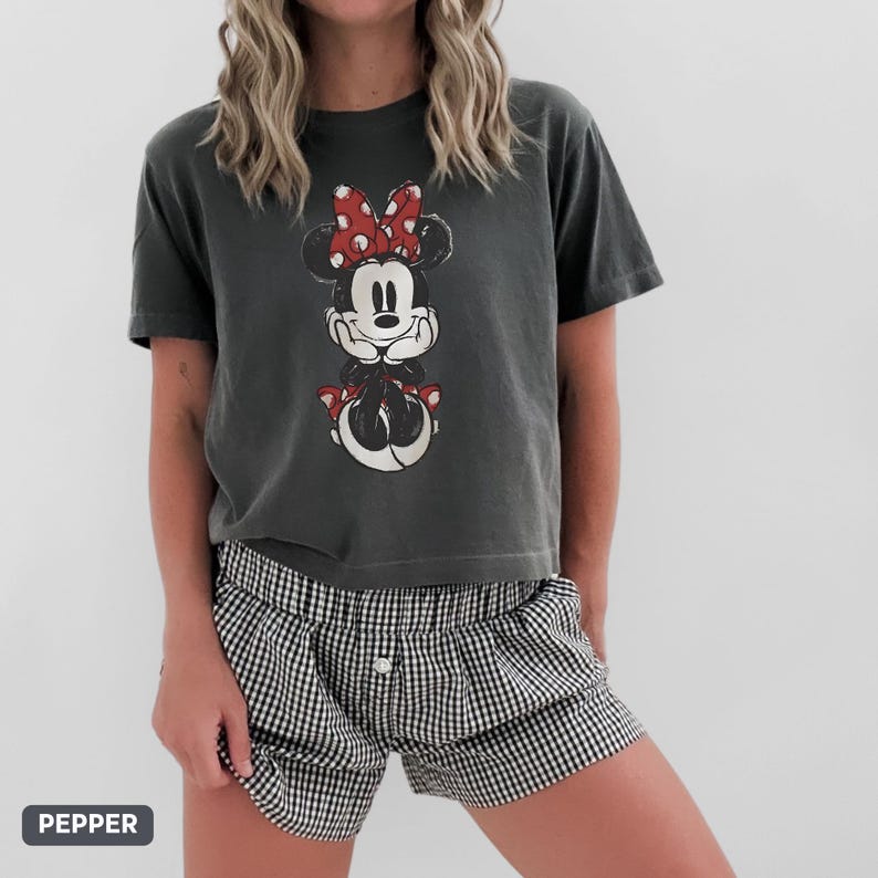 K&ouml;nnte beinhalten: Dunkelgraues T-Shirt mit einem verblassten Minnie Mouse-Motiv, roter Schleife und passenden Schuhen. Dazu eine schwarz-wei&szlig; karierte Shorts. Das Wort "PEPPER" steht in einem grauen Feld unten links.