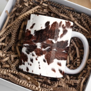 May include: White ceramic mug with a cowhide pattern in shades of brown, black, and white. The mug has a white handle and is presented in a white gift box with brown crinkle paper.