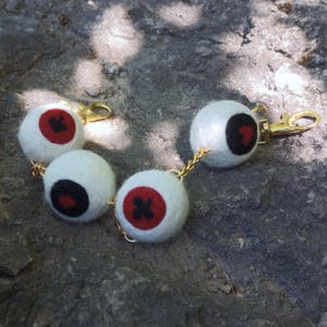 May include: Four white felt eyeball keychains with red and black iris details, connected by gold chains and clasps. The keychains are displayed on a gray stone surface, showcasing a unique and quirky accessory.