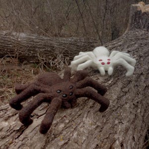May include: Two felted spider toys, one brown and one white, with red eyes, are posed on a tree trunk.