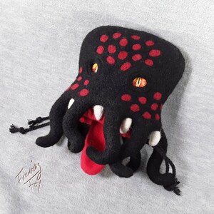 以下が含まれることがあります： 赤い斑点と黄色い目をしたタコの形をした黒いフェルト製のサイコロバッグ。バッグは口から赤いフェルトの舌が出ています。