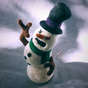Puede incluir: Un muñeco de nieve de fieltro blanco con un sombrero de copa negro, una bufanda verde y brazos marrones. El muñeco de nieve tiene una cruz negra en la cara y una nariz roja y naranja.