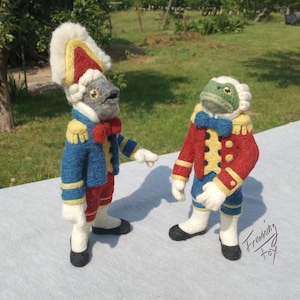 May include: Two felt figures wearing red and blue military uniforms with yellow accents. The figure on the left has a gray face and a white hat with a red and yellow trim. The figure on the right has a green frog face and a white hat with a red and yellow trim. The figures are standing on a white surface.