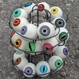 Könnte beinhalten: Ein dreistöckiger Metallständer zeigt eine Sammlung von weißen Filzkugeln, die jeweils mit einem anderen bunten Augenmotiv verziert sind. Die Augen sind mit verschiedenen Mustern bestickt, darunter Sterne, Zickzacklinien und Spiralen.