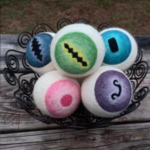 May include: Six white wool dryer balls with different colored designs. The designs include a green ball with a black zig-zag, a blue ball with a black rectangle, a purple ball with a black squiggle, a pink ball with a red dot, and two balls with black designs.