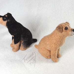 May include: Two needle felted dog figurines. The dog on the left is black with brown markings and is sitting with its head tilted up. The dog on the right is light brown and is sitting with its head tilted to the right. The dogs are both sitting on a white surface.