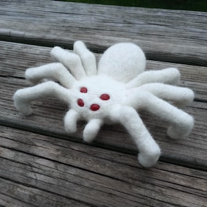 May include: A white felt spider with eight legs and red eyes rests on a wooden surface. The spider's body is round, and its legs are slightly curved. The red eyes provide a striking contrast to the white body.