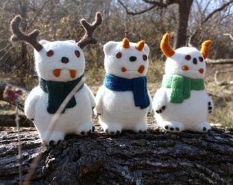May include: Three handmade white felt monster dolls with brown antlers, orange horns, and orange horns. Each doll has a different colored scarf: dark green, blue, and light green. The dolls are standing on a log.