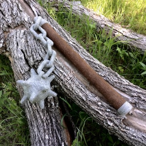 Op de afbeelding: Een handgemaakte viltknots met een bruine steel en een grijze, stekelige kop, verbonden door een grijze vilten ketting. De knots ligt op een verweerde boomstam in een natuurlijke omgeving.