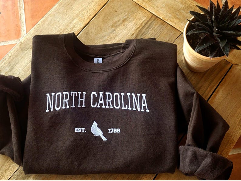 Embroidered maroon sweatshirt with “North Carolina” in bold white letters, featuring below a smaller text with year of establishment and a Northern Cardinal silhouette.