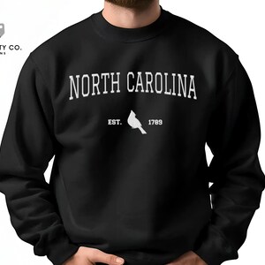 May include: Black sweatshirt featuring the text "NORTH CAROLINA" in white, arched across the chest. Below the text is a white cardinal and the text "EST. 1789". The sweatshirt is a crew neck style.