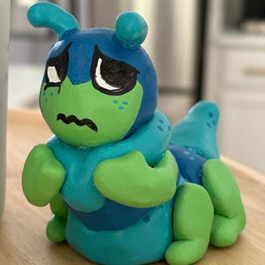 May include: A hand-painted, ceramic caterpillar figurine in shades of blue and green. The caterpillar has large, expressive eyes, a sad mouth, and small antennae. The figurine is sitting on a wooden surface.