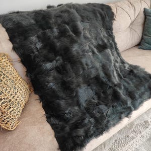 May include: A dark gray faux fur throw blanket draped over a beige sofa. The blanket has a plush, textured appearance. A woven decorative pillow and a teal pillow are also visible.