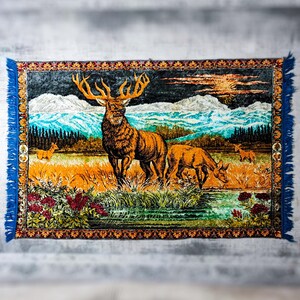 Pode incluir: Uma tapeçaria retangular tecida apresentando um grande veado com chifres proeminentes num prado dourado. A cena inclui um veado mais pequeno, uma cordilheira distante, uma floresta escura e um céu ao pôr do sol, com uma borda floral.