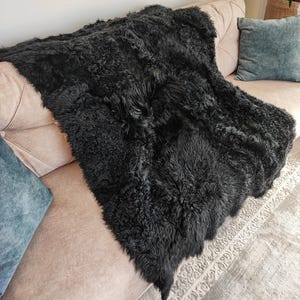 May include: A large, black sheepskin rug draped over a beige sofa. The rug has a thick, textured appearance and covers most of the sofa's surface. A teal pillow is visible on the sofa, and a patterned rug is partially visible on the floor.