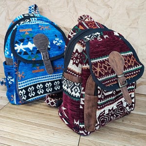 May include: Two patterned backpacks with a geometric design. One backpack is blue with white and orange accents, the other is red with white and green accents. Both backpacks have a brown leather flap with a buckle.