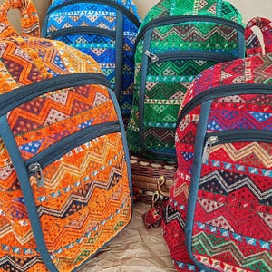 Op de afbeelding: Een verzameling kleurrijke rugzakken met een geometrisch patroon. De rugzakken zijn in tinten van oranje, blauw, groen en rood, met donkerblauwe ritsen en randen. De tassen hebben een afgeronde bovenkant en een voorvak met rits.