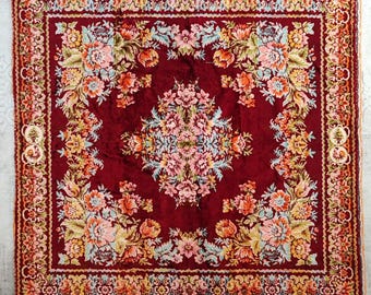 May include: A square, burgundy-colored rug with a floral pattern. The rug features a central floral design in shades of pink, peach, and yellow, surrounded by a detailed border. The rug's edges are finished with a decorative trim.