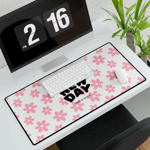 Peut inclure: Tapis de bureau blanc avec bordure noire, orné d'un motif floral rose et des mots "NEW DAY RISE N' SHINE" en noir. Un clavier et une souris blancs sont posés sur le tapis. Un écran d'ordinateur est visible en arrière-plan.