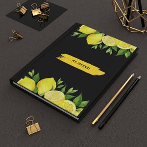 Peut inclure: Journal noir avec un coup de pinceau jaune portant l'inscription "MY JOURNAL". Le journal est orné de citrons à l'aquarelle et de feuilles vertes. Des crayons dorés et noirs et des pinces à reliure dorées sont également visibles.