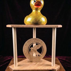 May include: A gold glitter duck trophy with a red beak and black eyes sits atop a wooden stand. The stand features a wooden medallion with the text "LET' ER FLY ARCHERY" and a bird graphic, with the text "1st Place Battle of the Ages".