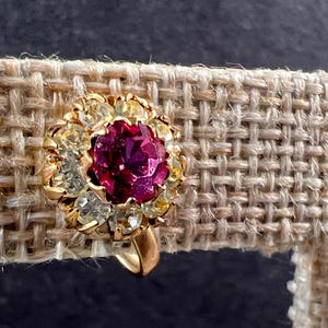 May include: A gold-coloured clip-on earring featuring a central, deep pink gemstone encircled by smaller clear and yellow stones. The earring is set against a textured, beige fabric background.