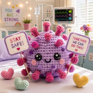 May include: A round, purple crocheted plush toy with a smiling face, large black eyes, and pink cheeks. It features protruding pink spikes and holds two white rectangular signs with the text STAY SAFE! and TOGETHER WE CAN DO THIS!.