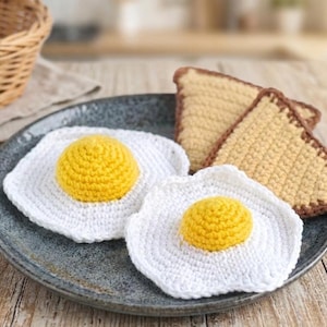 May include: A plate holds two crocheted fried egg trivets with yellow yolks and white whites, and two crocheted toast slices with brown edges. The items are handmade and sit on a gray plate.