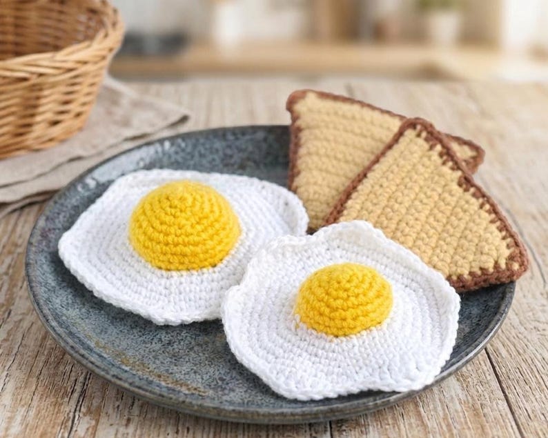 May include: A plate holds two crocheted fried egg trivets with yellow yolks and white whites, and two crocheted toast slices with brown edges. The items are handmade and sit on a gray plate.