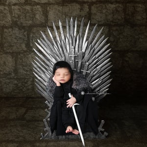 Könnte beinhalten: Ein Baby in einem schwarzen Kapuzengewand mit Pelzkragen sitzt auf einem silbernen Thron und hält ein Schwert in der Hand. Der Thron ist aus Metall und hat ein scharfes, spitzes Design.