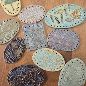 May include: A collection of ten ceramic pieces in various shapes, including ovals and a rectangle, each featuring pre-drilled holes along the edges. The designs include embossed patterns of fish, dragonflies, floral motifs, and wavy textures in colors like blue, brown, green, and tan.
